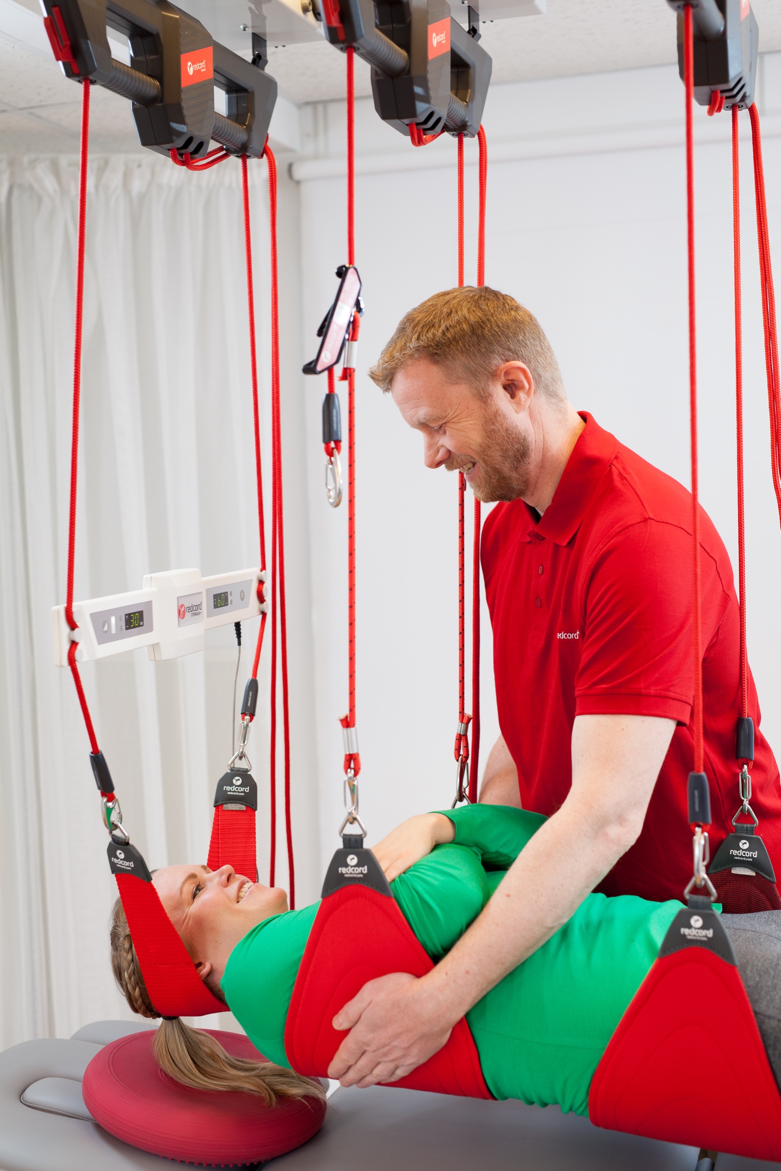 A Redcord practitioner guiding a smiling patient through a supported pelvic-lift exercise on the Redcord Workstation